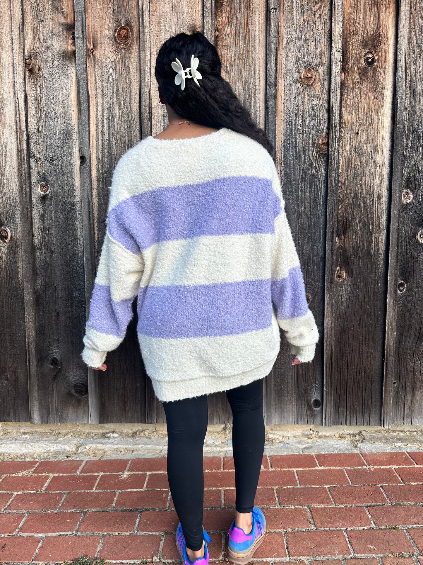 A woman wearing an oversized striped henley sweater in lavender and white colors with functional buttons, ribbed trim, and a subtle hi-lo fit.
