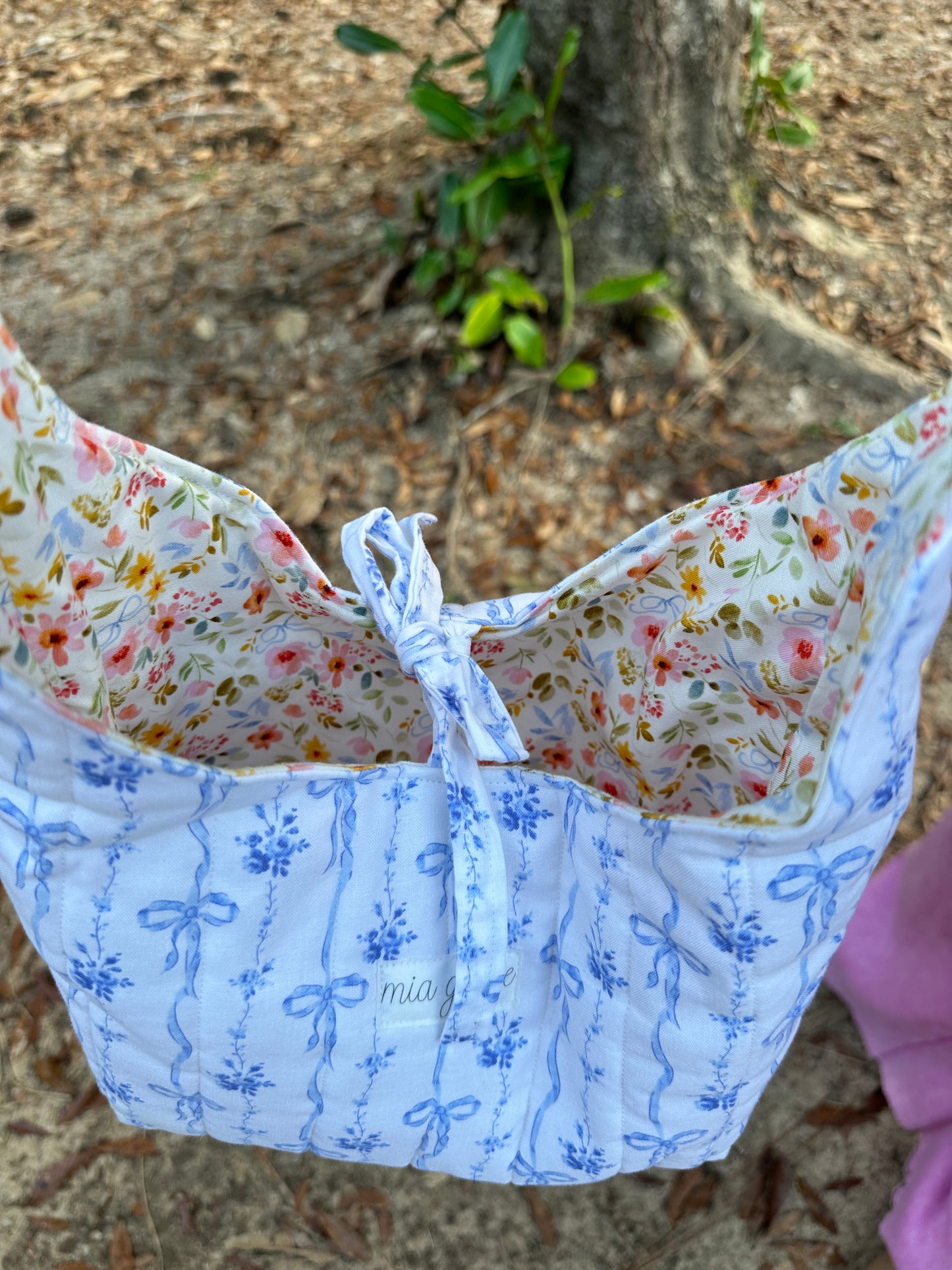 person wearing a blue bows and floral fabric shoulder bag with a tie closure and a Mia grace brand label.  inner fabric is florals with blue bows on a cream colored background. 