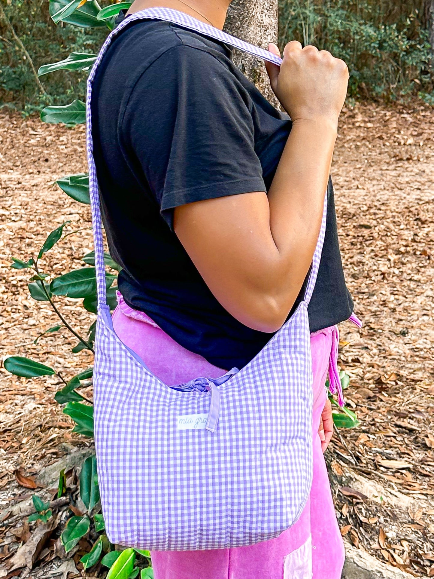 woman wearing a purple gingham shoulder bag with a Mia Grace brand label and a purple tie closure to match the purple inner fabric. 