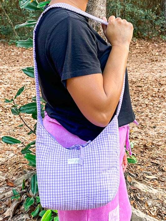 woman wearing a purple gingham shoulder bag with a Mia Grace brand label and a purple tie closure to match the purple inner fabric. 