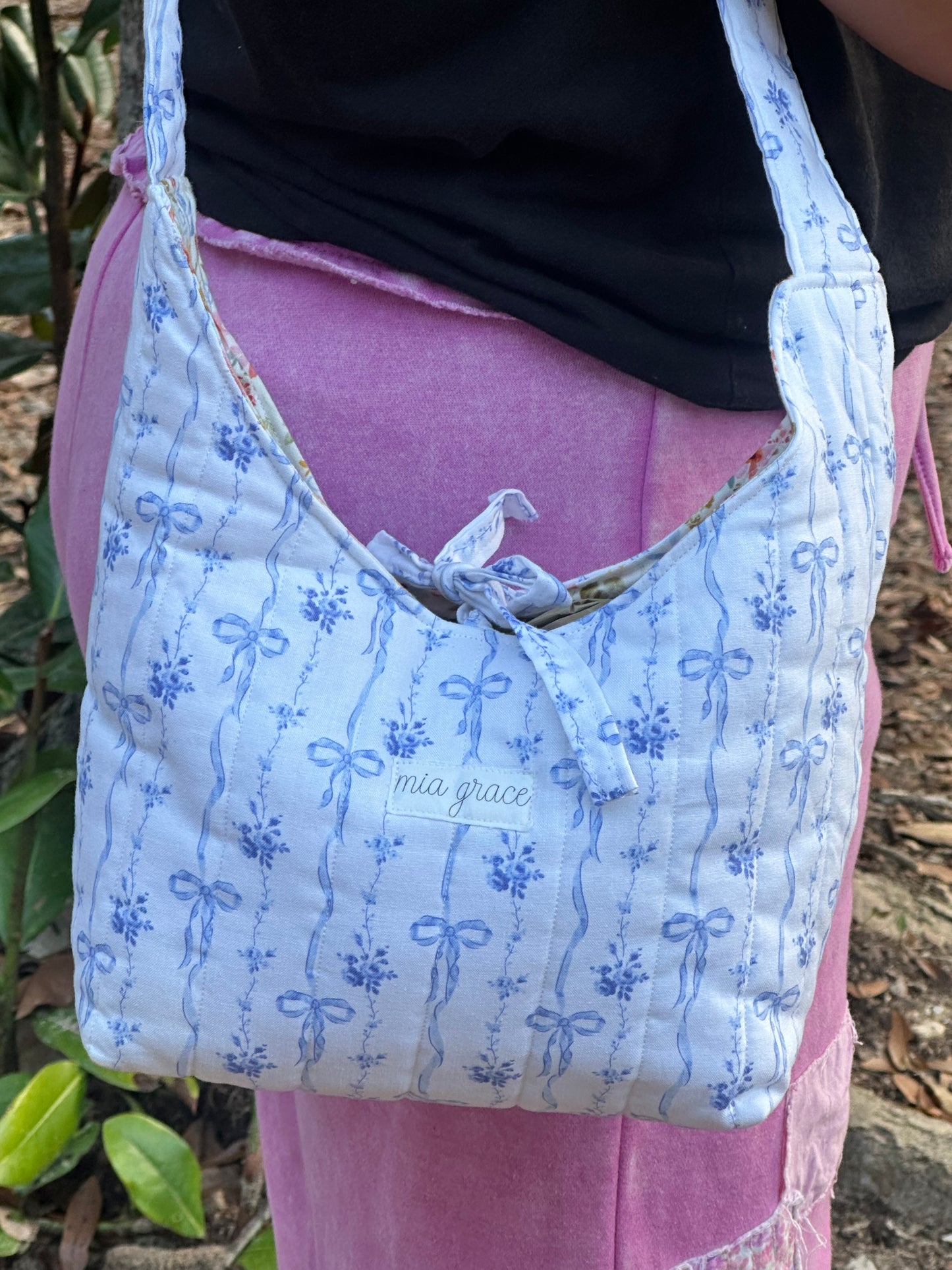person wearing a blue bows and floral fabric shoulder bag with a tie closure and a Mia grace brand label.  inner fabric is florals with blue bows on a cream colored background. 