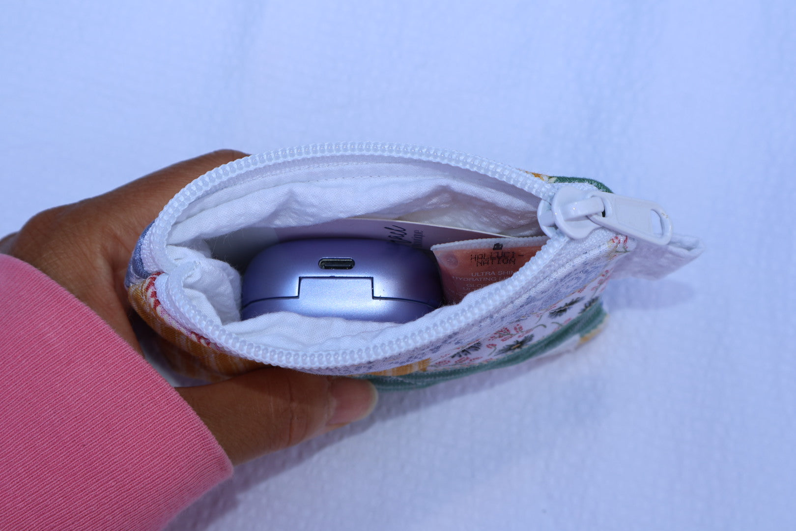 close up of a small pouch with white inner fabric and a zipper showcasing how many items that it can hold. Displays earbuds, mini lip gloss, and a card that is not included with the wallet. 