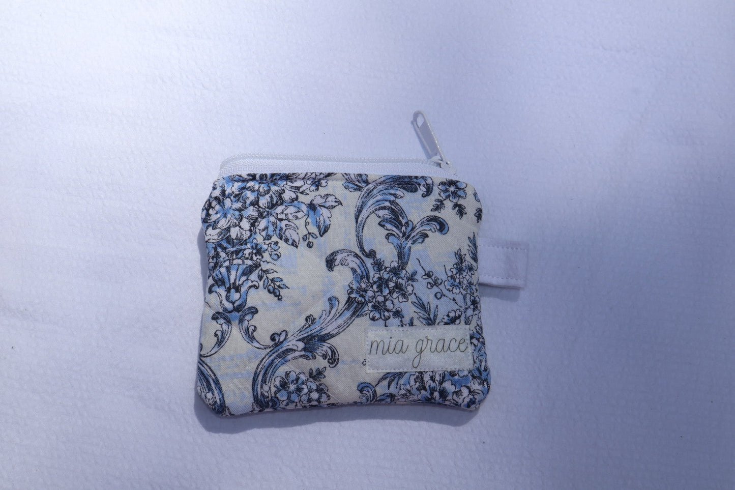 Small  blue and white floral wallet sized pouch with a 'mia grace' brand label, zipper closure, and a hoop for a key ring to be attached. 