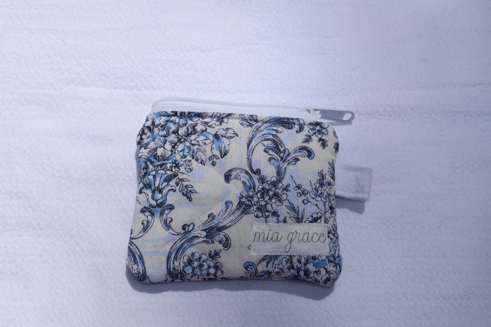 Small  blue and white floral wallet sized pouch with a 'mia grace' brand label, zipper closure, and a hoop for a key ring to be attached. 