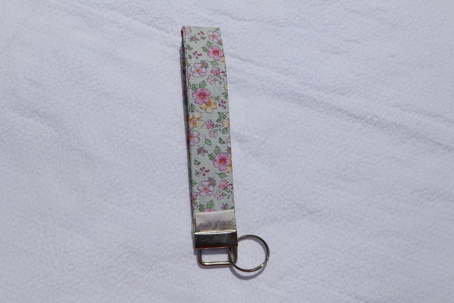 Fabric key fob with a mint, pink, and yellow floral  patten with butterflies and a Mia grace brand label and a key ring