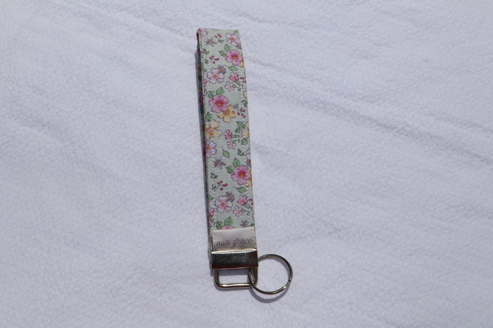 Fabric key fob with a mint, pink, and yellow floral  patten with butterflies and a Mia grace brand label and a key ring
