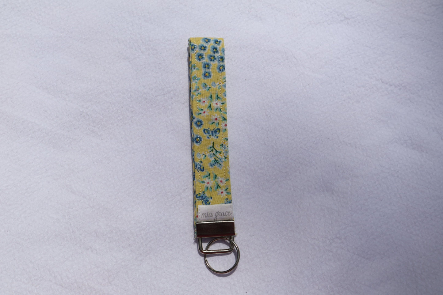 Fabric key fob with a yellow and blue floral  patten with butterflies and a Mia grace brand label and a key ring