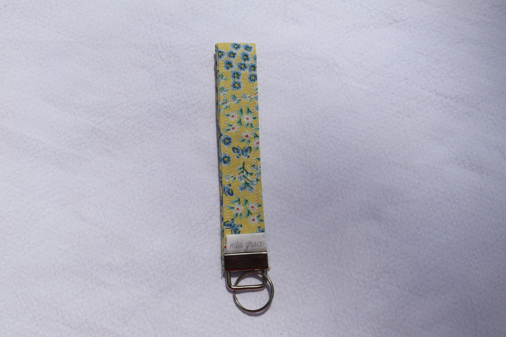 Fabric key fob with a yellow and blue floral  patten with butterflies and a Mia grace brand label and a key ring