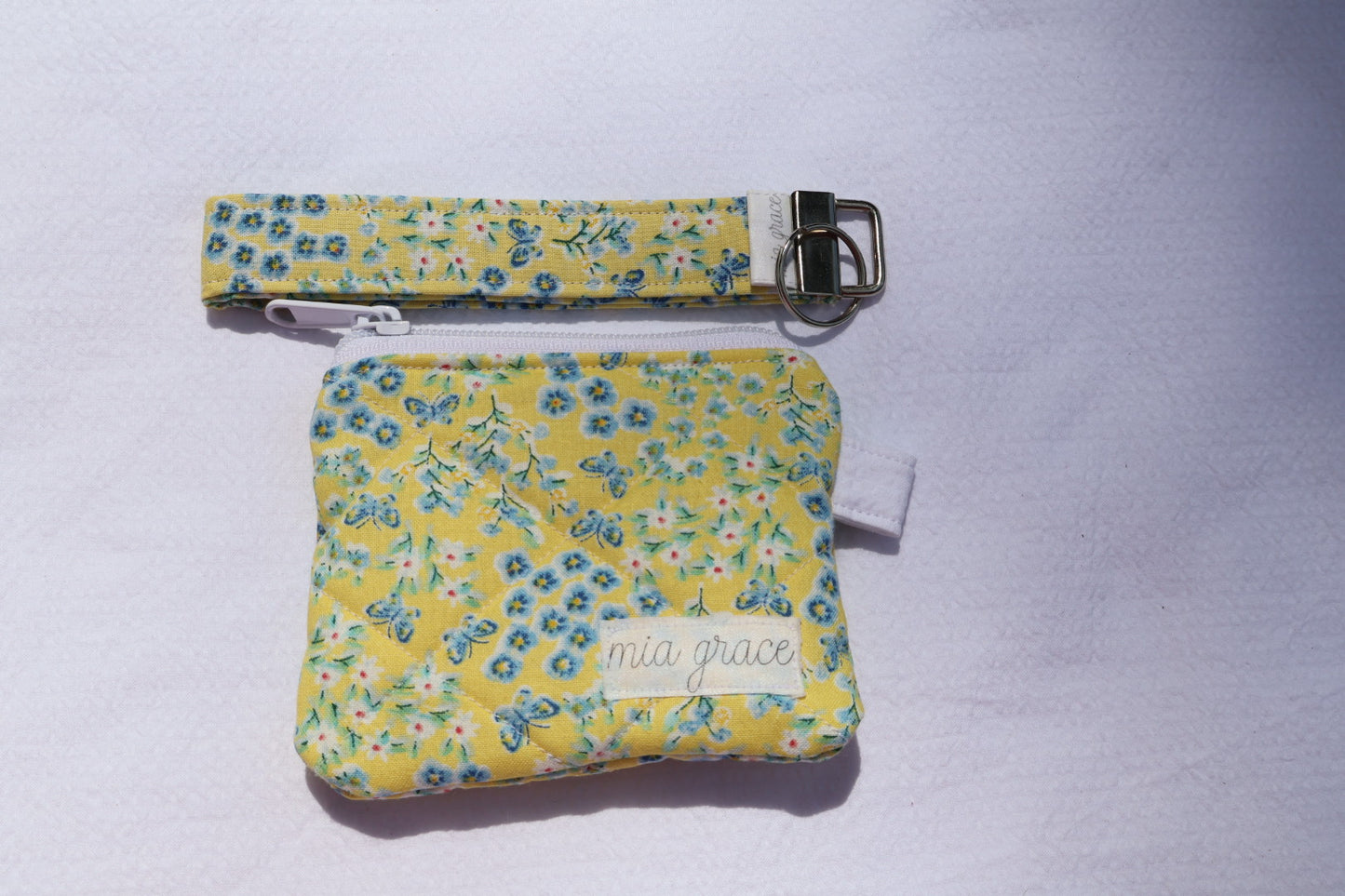 Fabric key fob with a yellow and blue floral  patten with butterflies and a Mia grace brand label and a key ring with a matching wallet that is sold separately. 