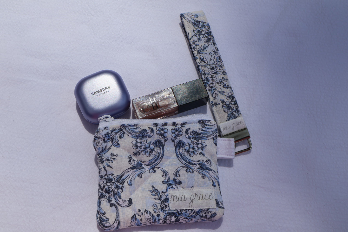 Small  blue and white floral wallet sized pouch with a 'mia grace' brand label, zipper closure, and a hoop for a key ring to be attached. 