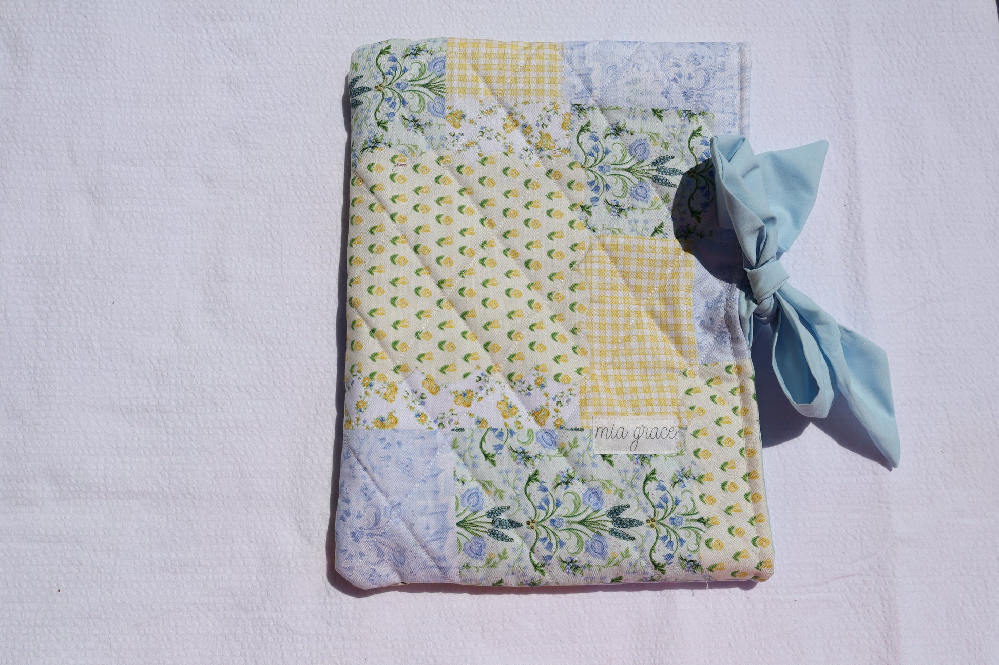 Quilted bible or book sleeve  with floral patterns in a patchwork print blue, yellow, and green. soft blue bow tie closure. Mia grace brand label on the front