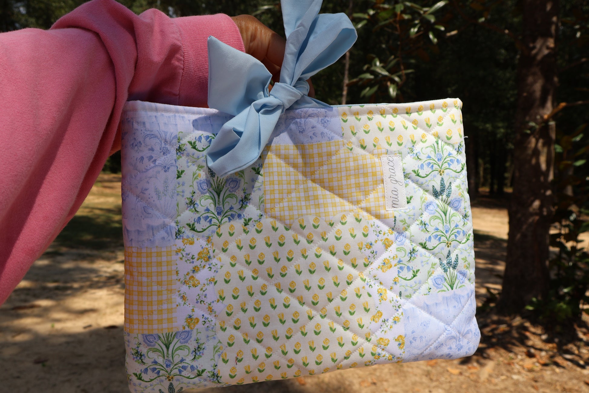 Quilted bible or book sleeve  with floral patterns in a patchwork print blue, yellow, and green. soft blue bow tie closure. Mia grace brand label on the front