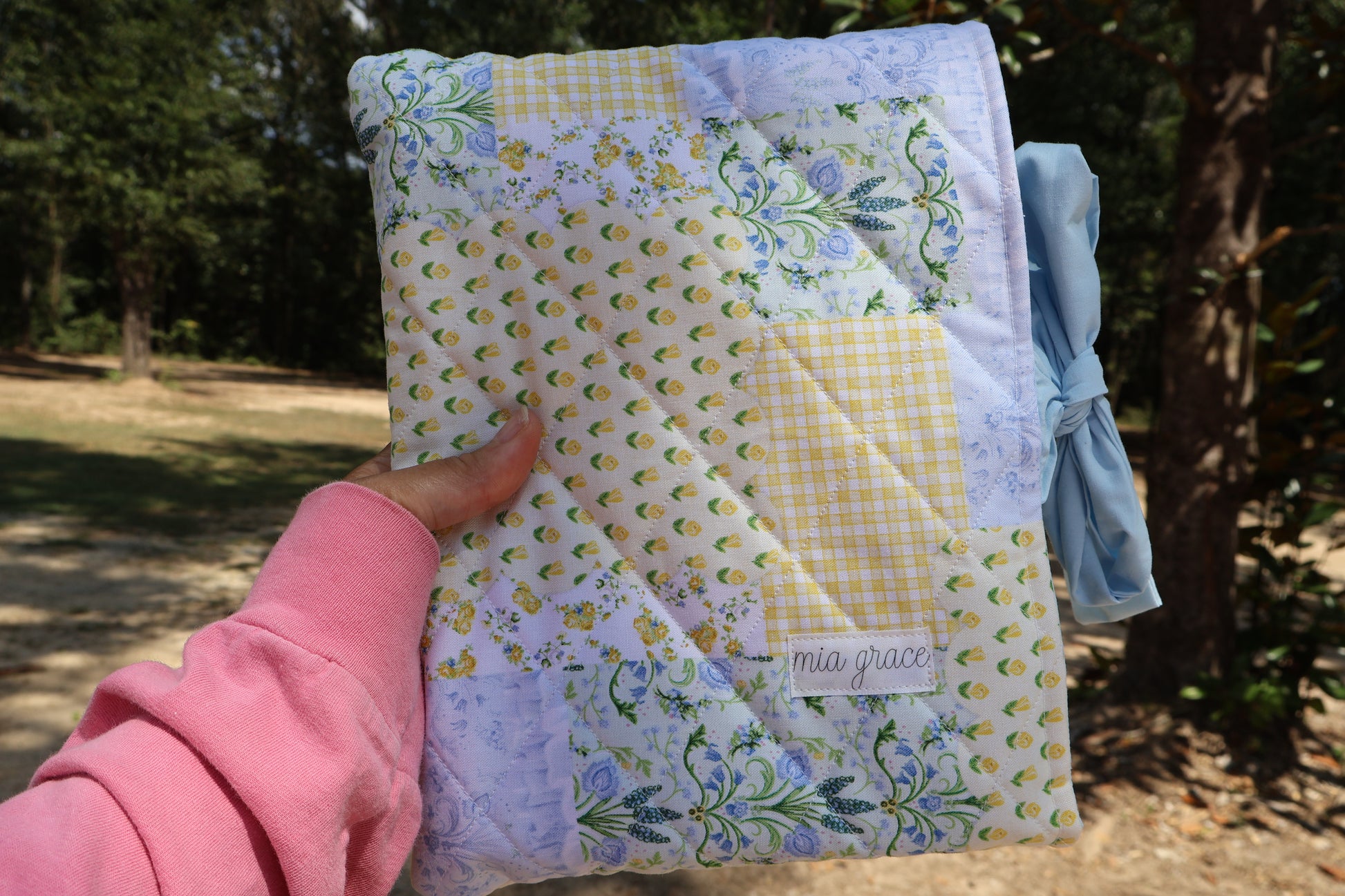 Quilted bible or book sleeve  with floral patterns in a patchwork print blue, yellow, and green. soft blue bow tie closure. Mia grace brand label on the front