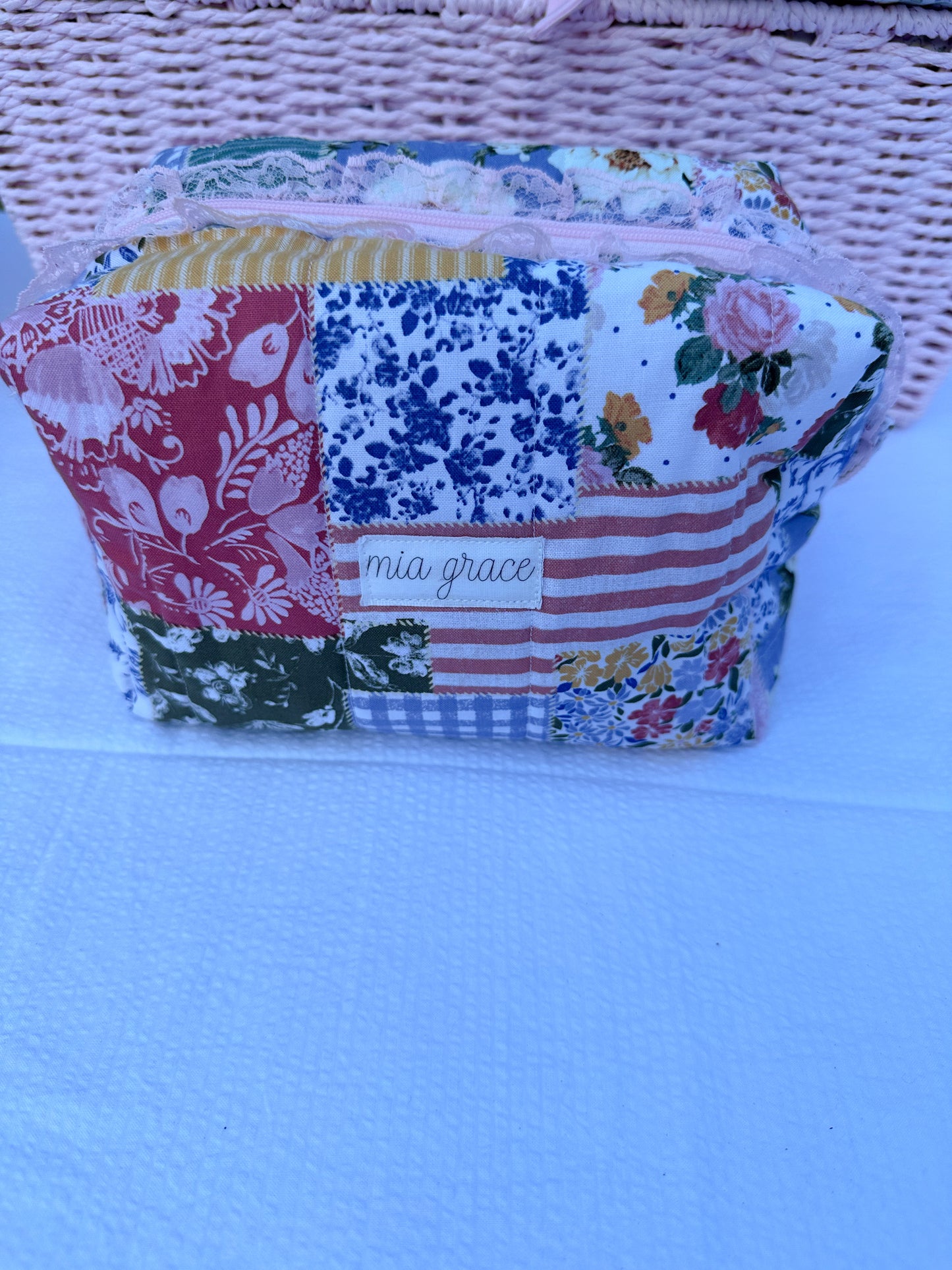 Patchwork-style fabric quilted boxy pouch with floral and gingham  patterns with a Mia grace brand label and pink zipper with pink lace trim and pull tabs.