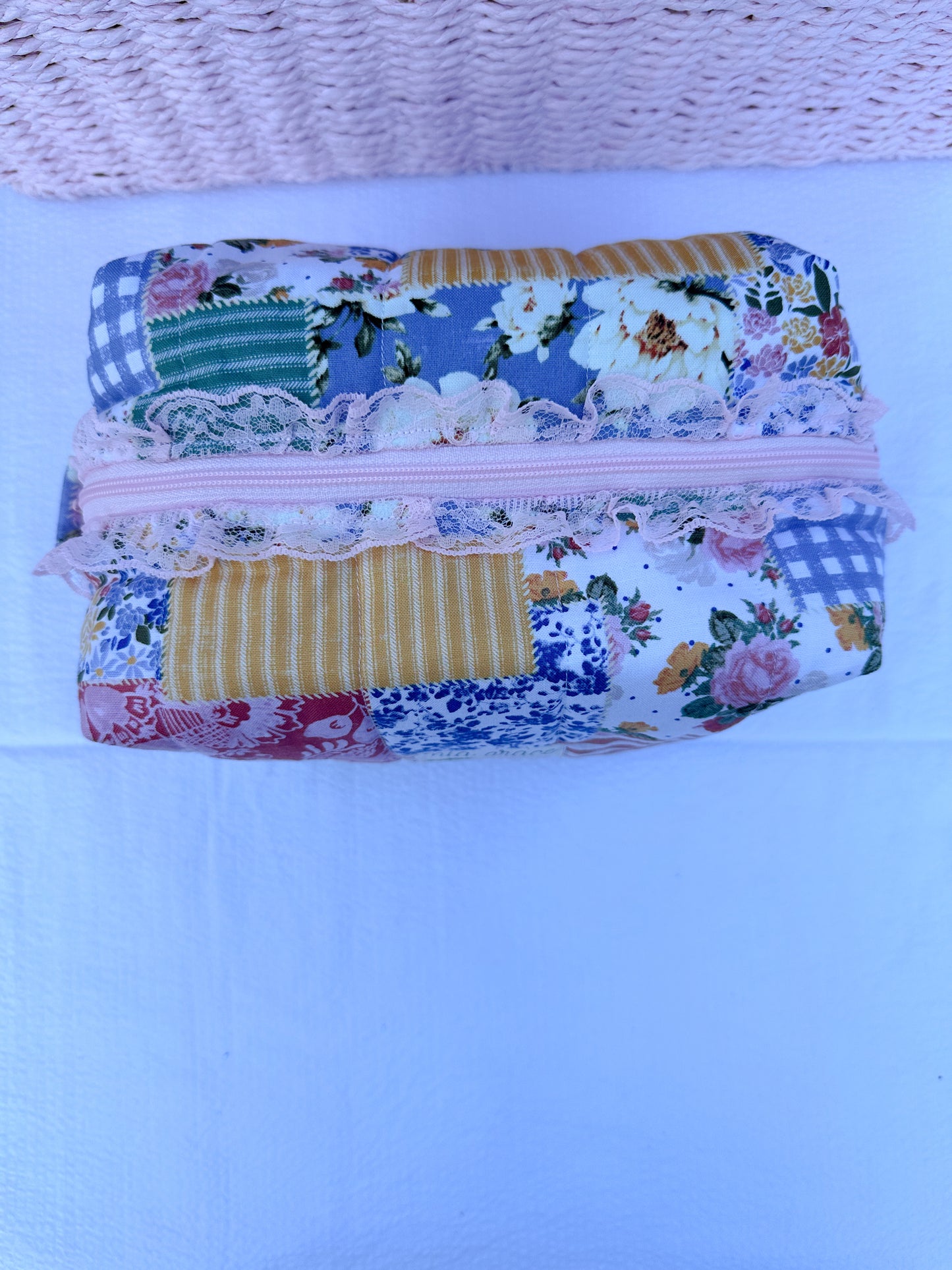 Patchwork-style fabric quilted boxy pouch with floral and gingham  patterns with a Mia grace brand label and pink zipper with pink lace trim and pull tabs.