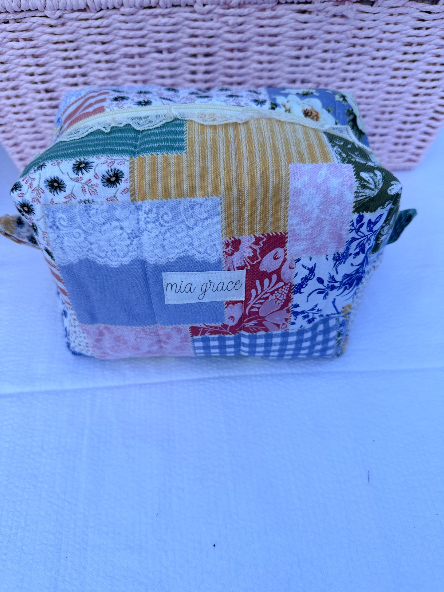Patchwork-style fabric quilted boxy pouch with floral and gingham  patterns with a Mia grace brand label and cream zipper with cream one sided lace trim and pull tabs.