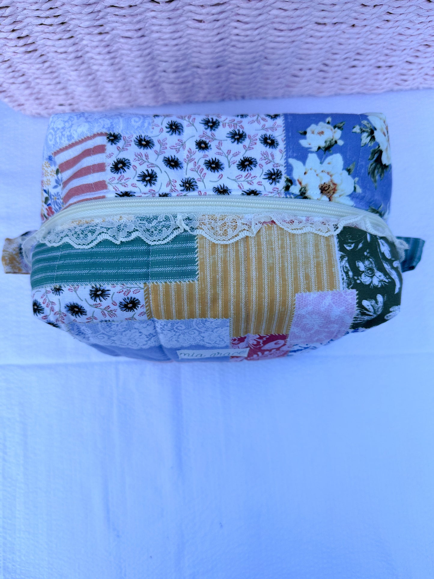 Patchwork-style fabric quilted boxy pouch with floral and gingham  patterns with a Mia grace brand label and cream zipper with cream one sided lace trim and pull tabs.