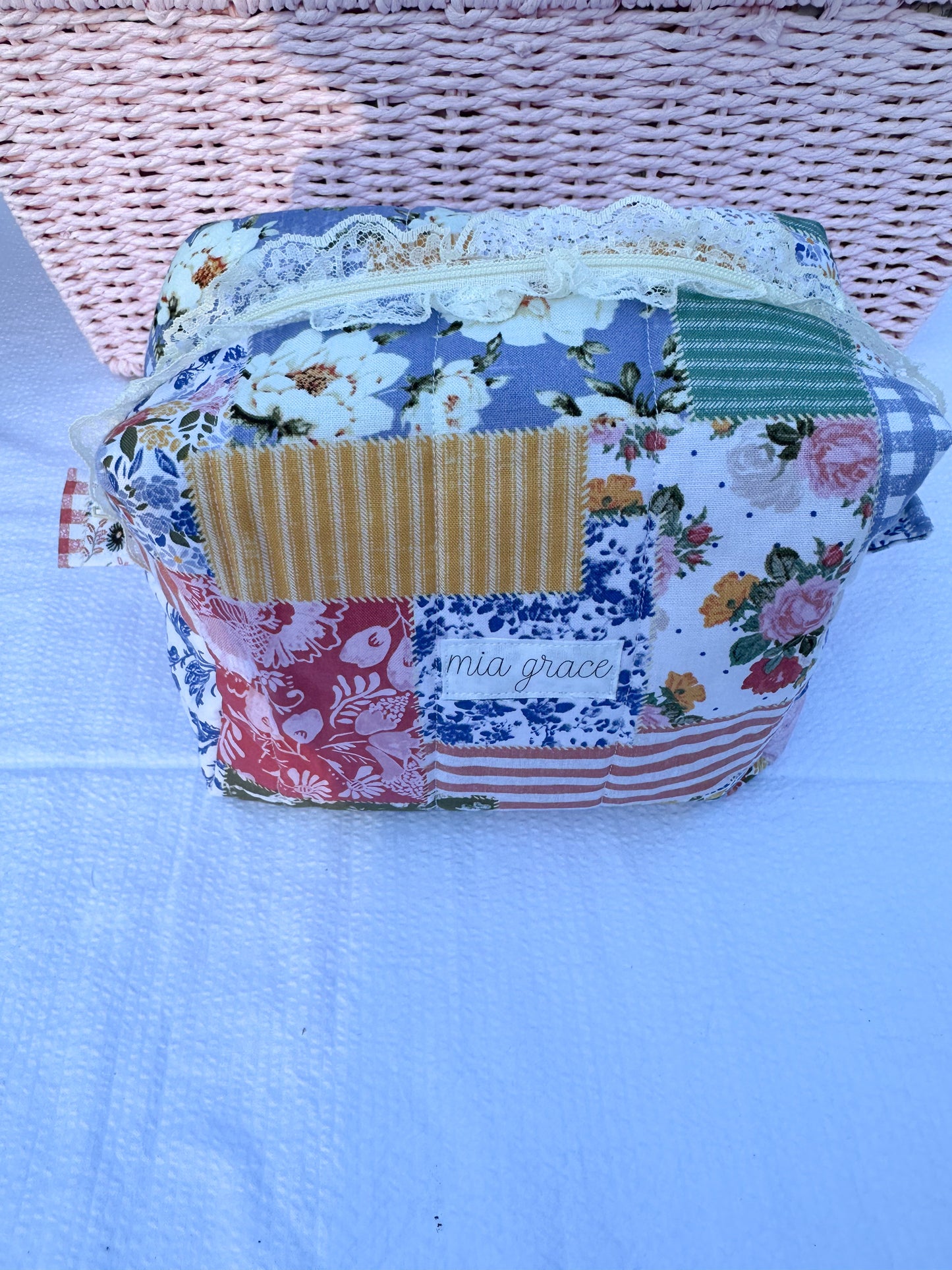 Patchwork-style fabric quilted boxy pouch with floral and gingham  patterns with a Mia grace brand label and cream zipper with cream  lace trim and pull tabs. 