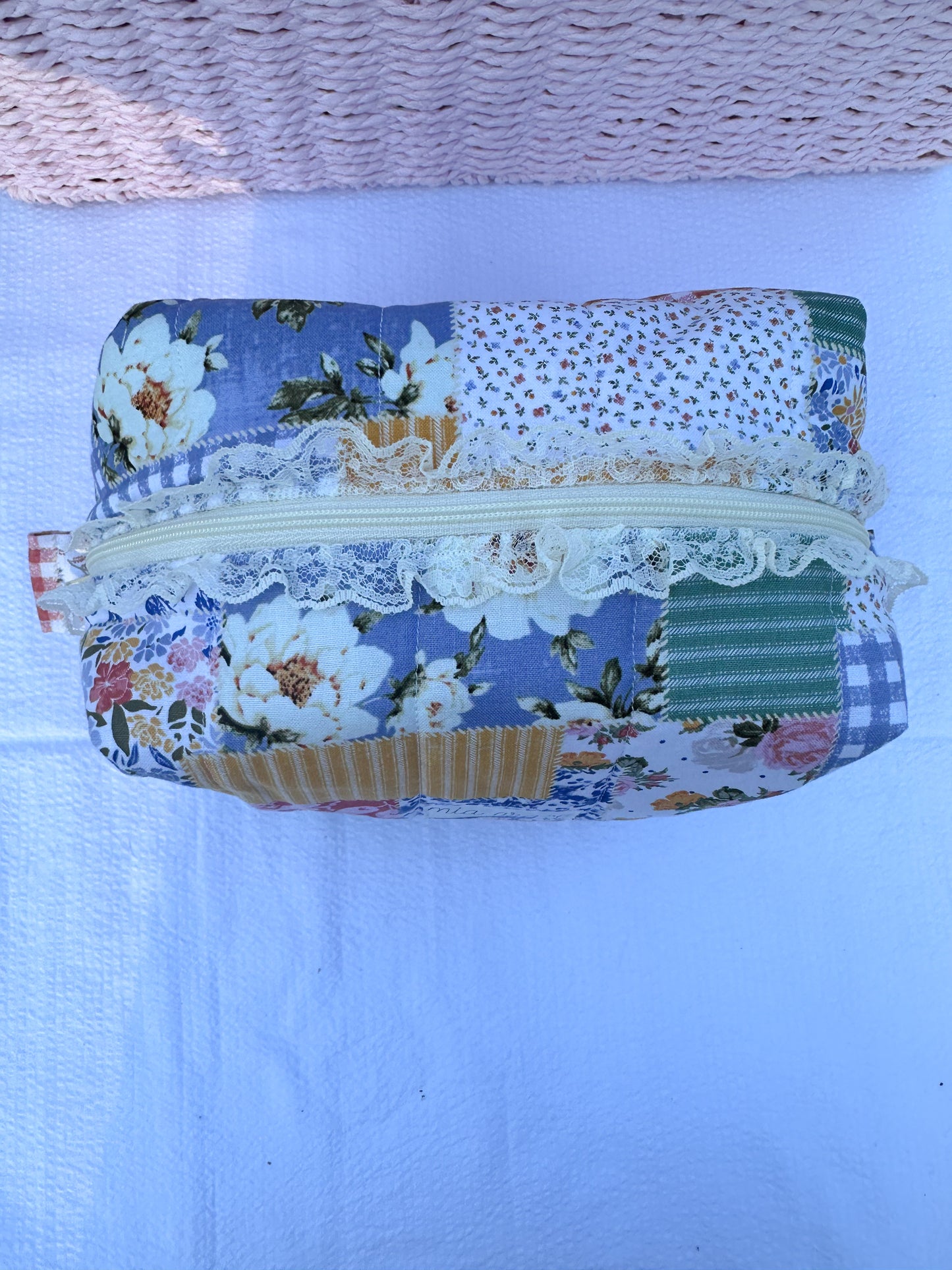 Patchwork-style fabric quilted boxy pouch with floral and gingham  patterns with a Mia grace brand label and cream zipper with cream  lace trim and pull tabs. 