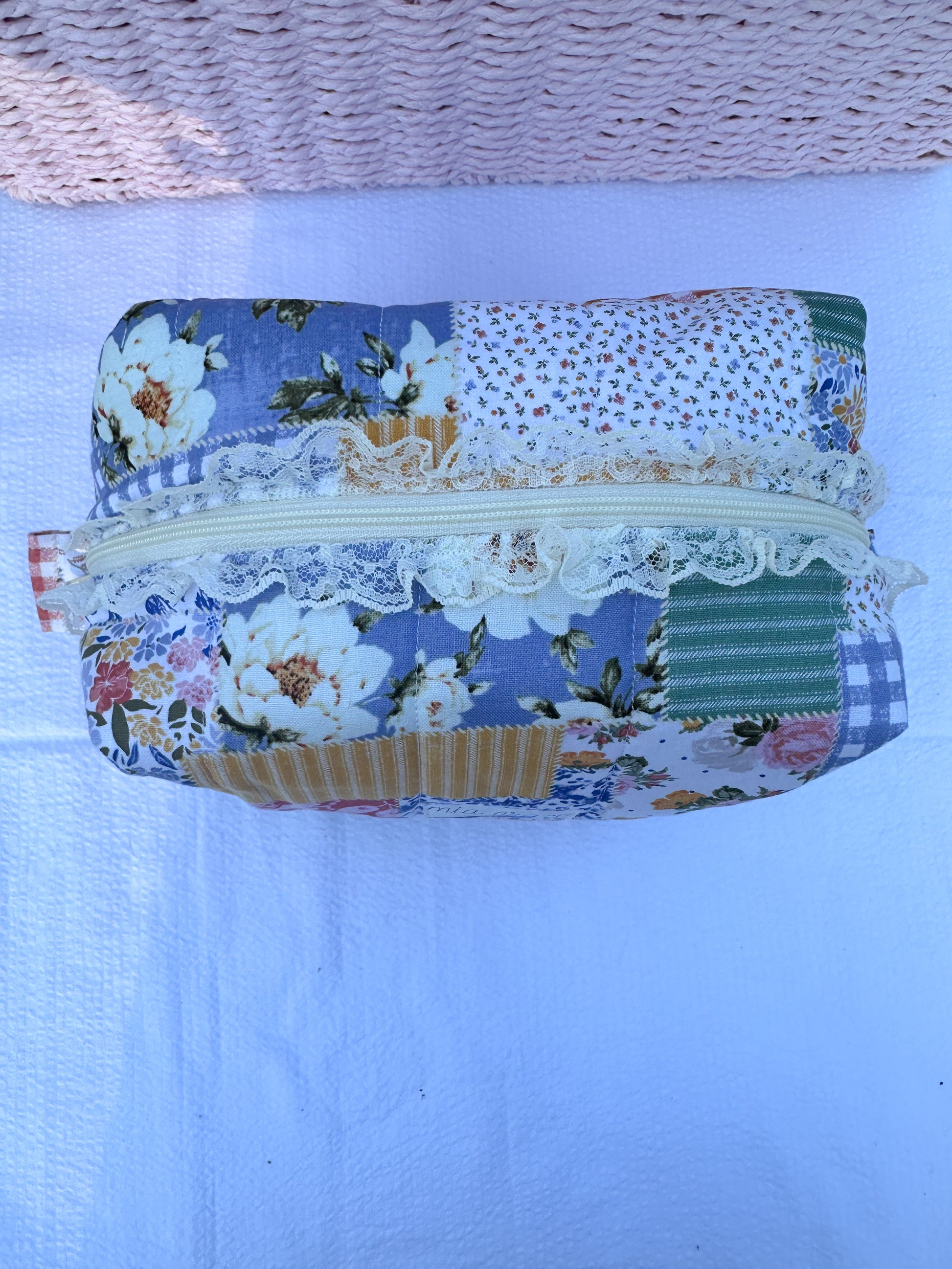 Patchwork-style fabric quilted boxy pouch with floral and gingham  patterns with a Mia grace brand label and cream zipper with cream  lace trim and pull tabs. 