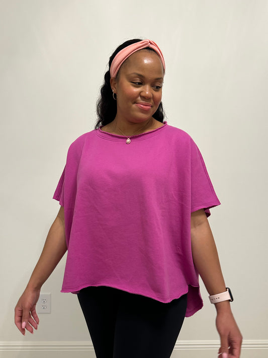 woman wearing an orchid color short sleeve dolman sleeve top that has a slight boatneck neckline and a hi-lo raw hem. 