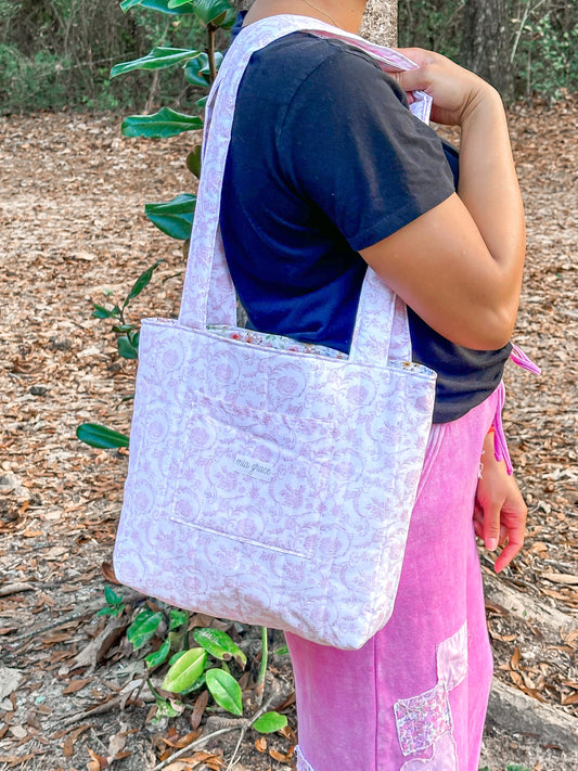 Person holding a pink and white floral paisley tote bag. Tote bag has a front pocket with a Mia grace brand label and an inner pocket with a bow label. inner fabric is a floral print with blue bows. 