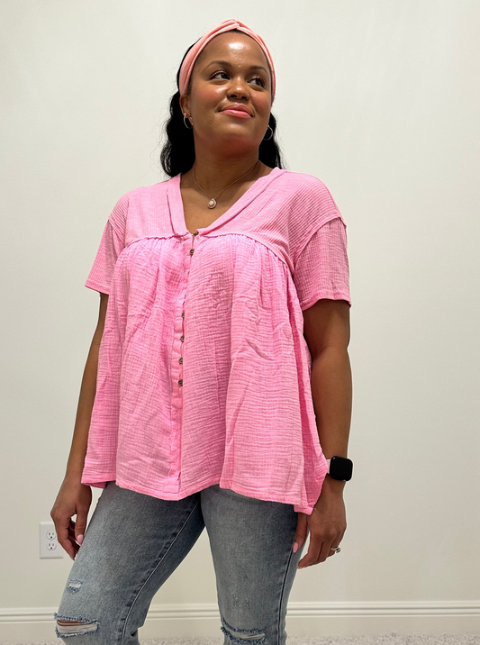 Person wearing a pink shirt that is a loose flowy babydoll style with buttons 2/3 down the front of the shirt with a side slit.