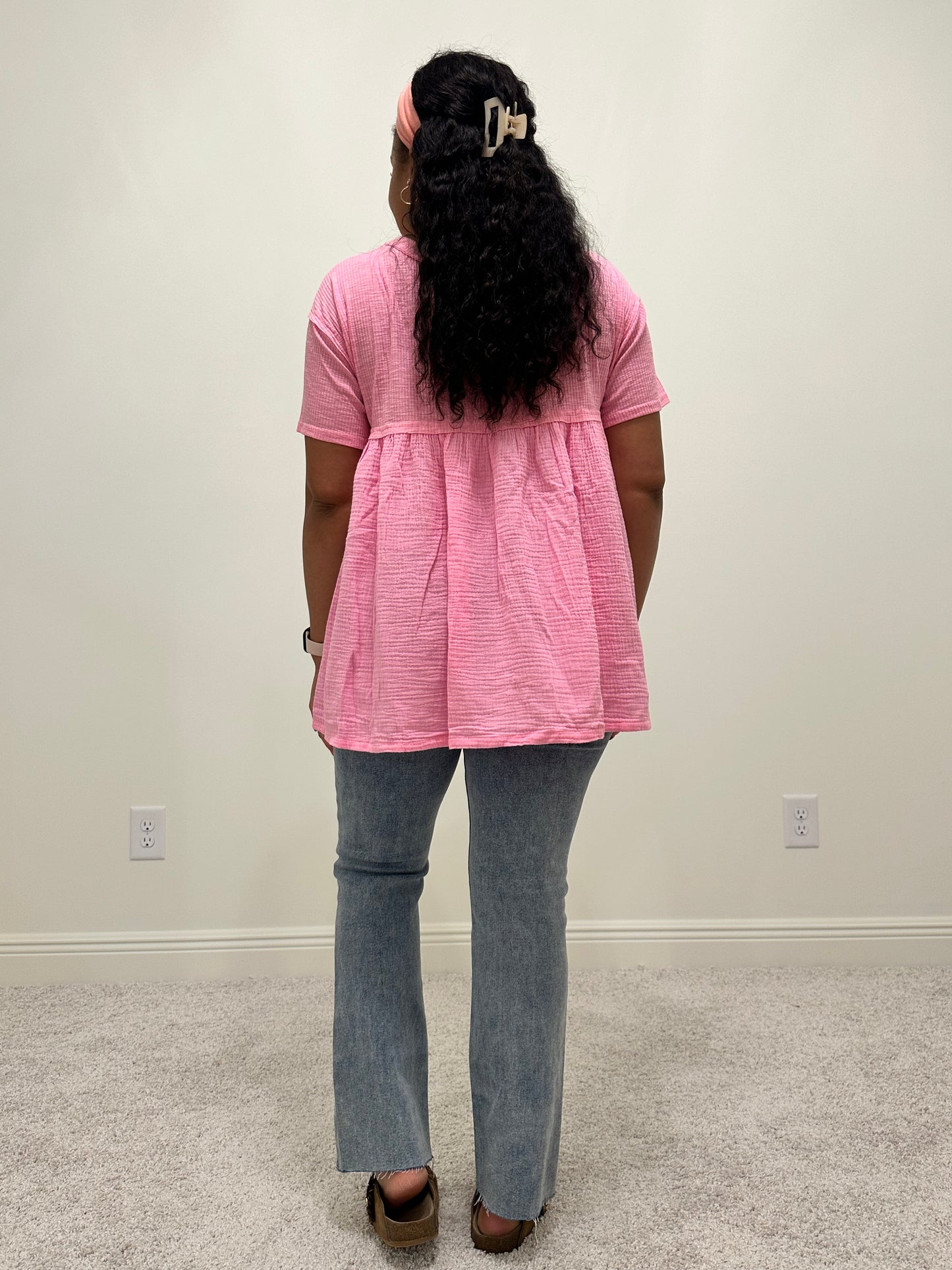 Person wearing a pink shirt that is a loose flowy babydoll style with buttons 2/3 down the front of the shirt with a side slit.