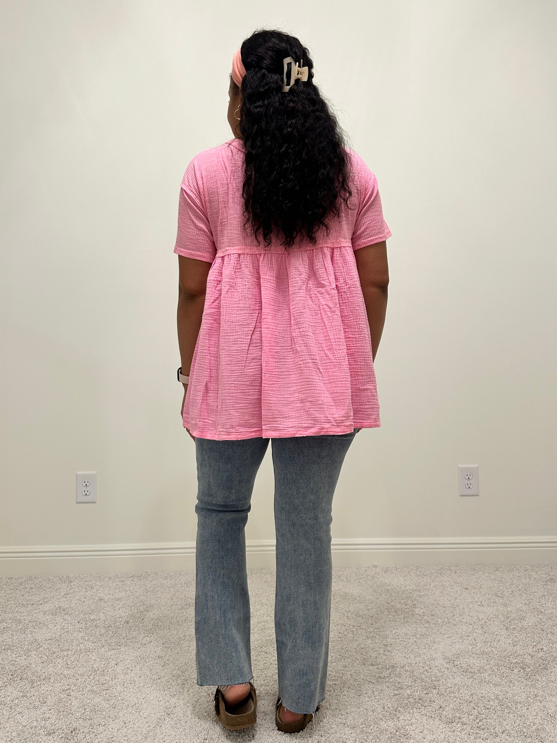 Person wearing a pink shirt that is a loose flowy babydoll style with buttons 2/3 down the front of the shirt with a side slit.